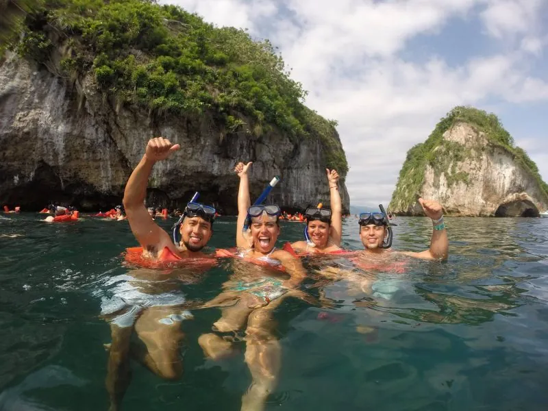 Four snorkelers in masks waving in the water below the cliffs of Los Arcos