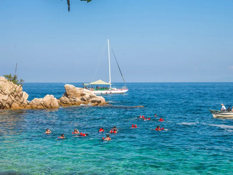 Snorkelers in orange vests near rocky islets with a sailboat anchored beyond