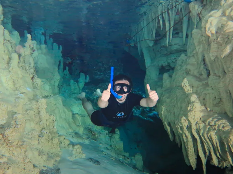 A snorkeler giving a thumbs up underwater between pale rock formations in a cenote