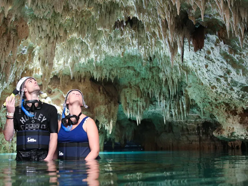 Two snorkelers in helmets standing in clear water under dense stalactites
