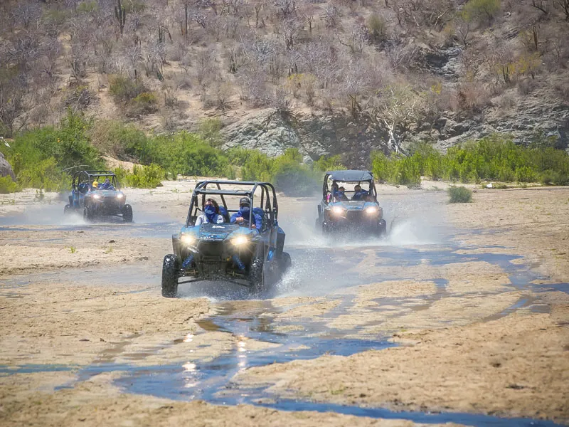 Three UTVs splashing across a shallow stream in a desert arroyo