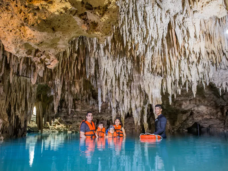 A family in life jackets standing in blue water under a ceiling of stalactites in a cenote