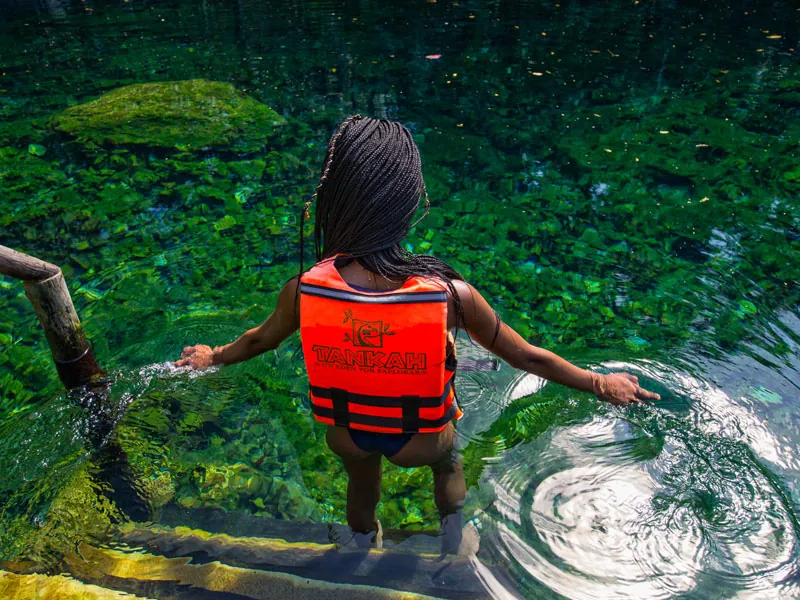 A woman in a Tankah life vest stepping down into clear green cenote water