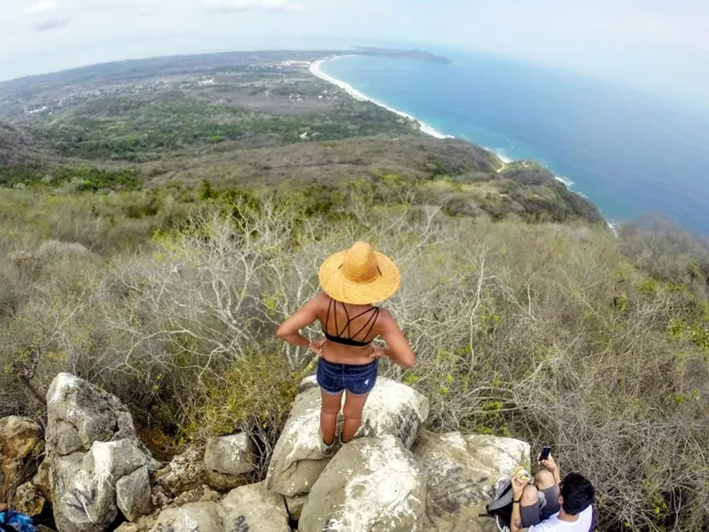 Hiker in a straw hat on the rocky summit of Cerro del Mono looking over the Punta de Mita coastline
