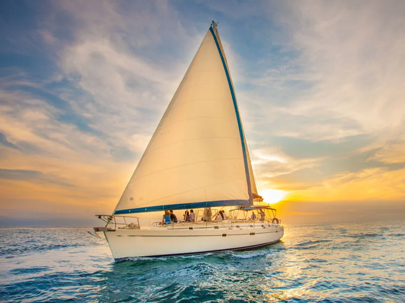 Sailing yacht under a white mainsail heading into a golden sunset with guests aboard