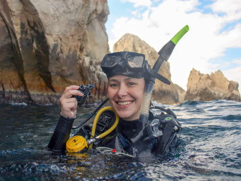 Smiling diver at the surface holding a camera, the Land’s End rocks behind