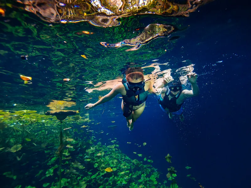 Two snorkelers in life vests photographed underwater in deep blue cenote water