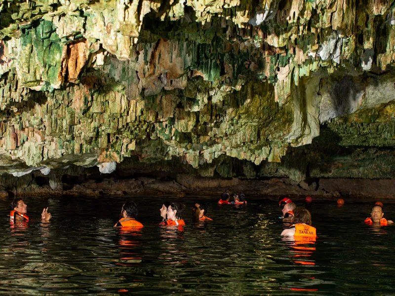 Swimmers in orange life vests in dark water under a cave ceiling of stalactites