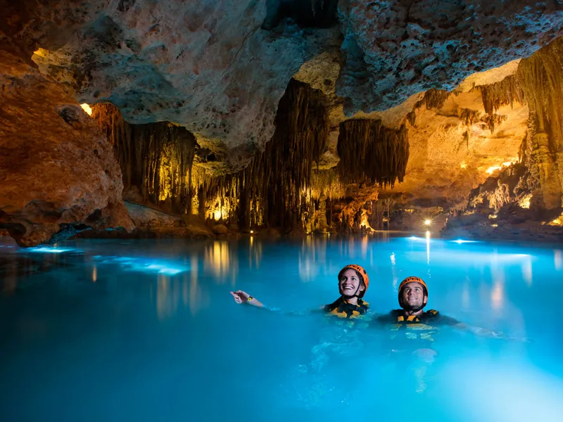 Two swimmers in helmets in glowing blue water inside a cave hung with lit stalactites