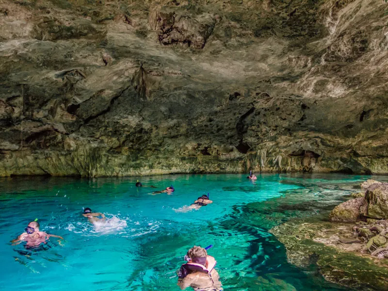 Snorkelers swimming in turquoise water beneath the rock ceiling of a cenote cave