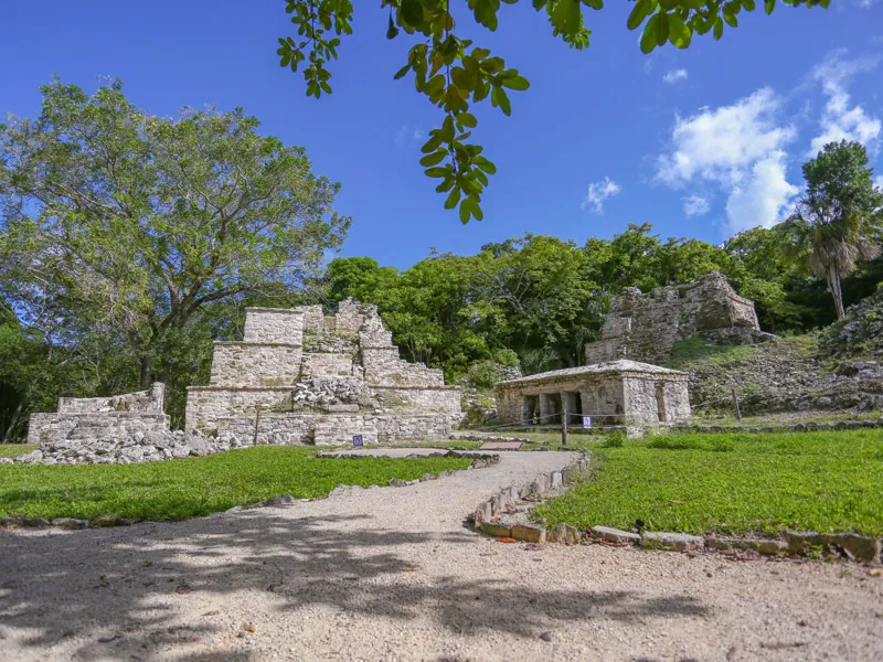 Low stone temples at Muyil beside a gravel path and green lawns under a blue sky