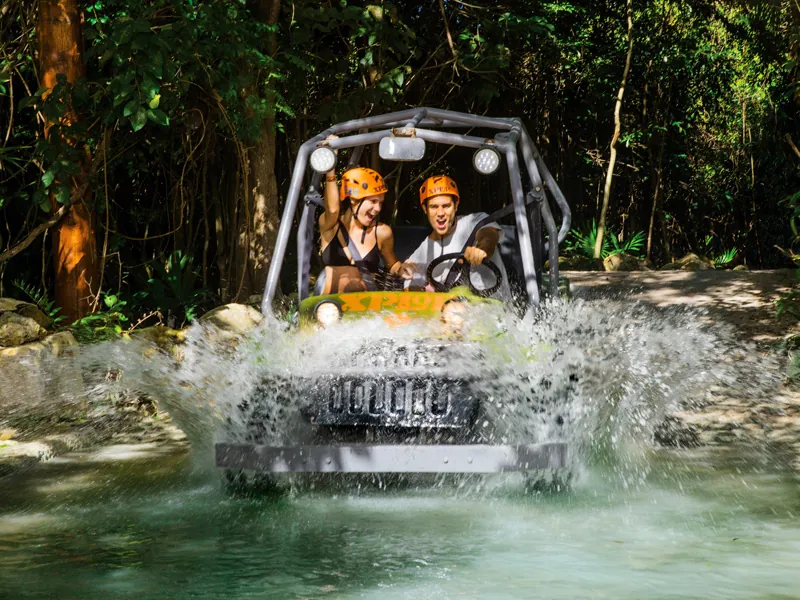 An amphibious vehicle with two helmeted guests splashes through water on a jungle track