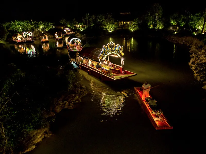 Illuminated trajineras move along a canal lined with lit trees at night