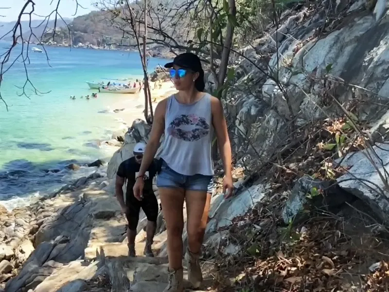 Hikers climbing a rocky coastal path with the beach and boats of the cove below