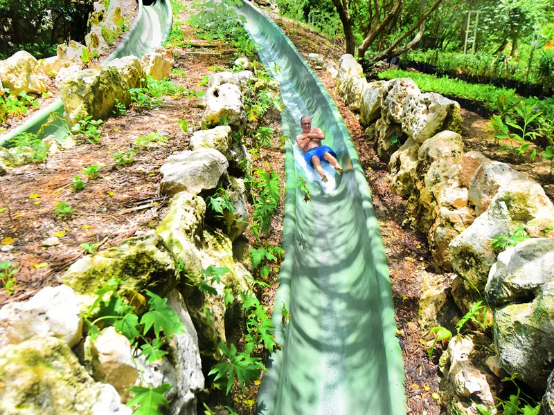 A guest rides a long green waterslide down a rocky garden slope at Xenses