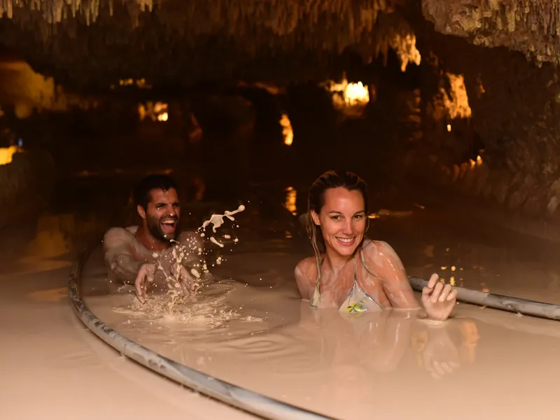 A smiling couple sit in a pool of pale mud inside a cave at Xenses