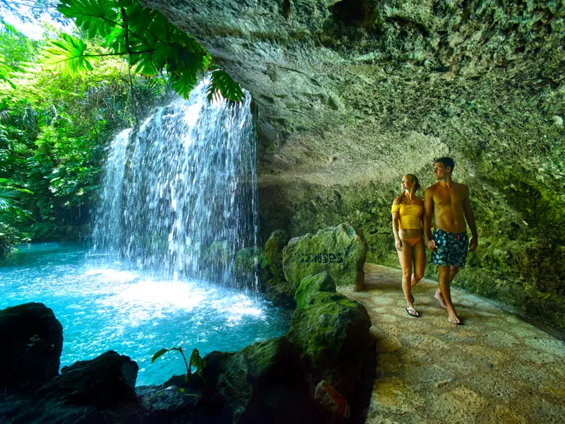 A couple walks a rocky path behind a waterfall pouring into a turquoise pool at Xenses
