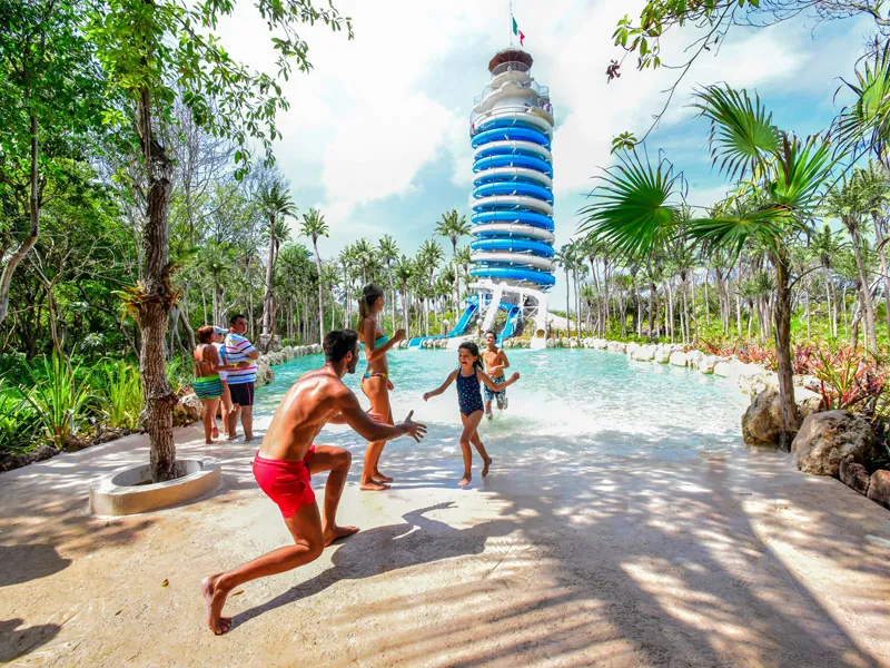 A family plays on a shaded sandy path in front of the blue-and-white lighthouse at Xel-Há