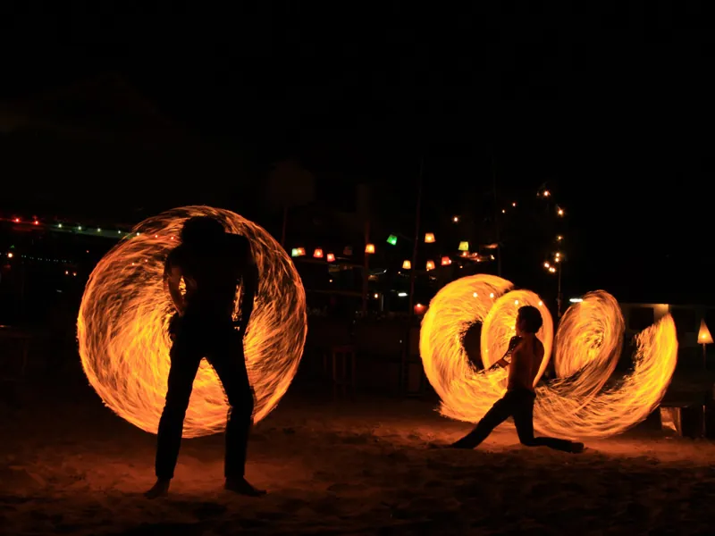 Three fire dancers spinning orange flame rings on a beach with lights behind