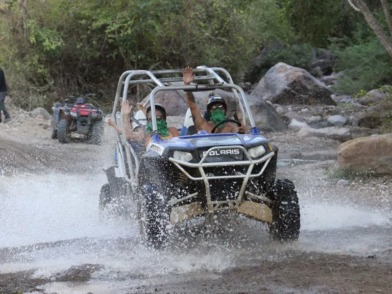 Guests raising their arms in a white RZR as it drives through a rocky river