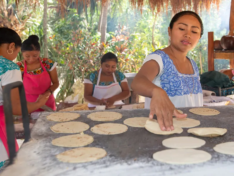 Maya women in embroidered blouses making corn tortillas on a large griddle