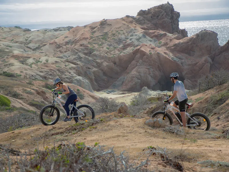 Two cyclists on electric bikes on a trail below eroded desert hills