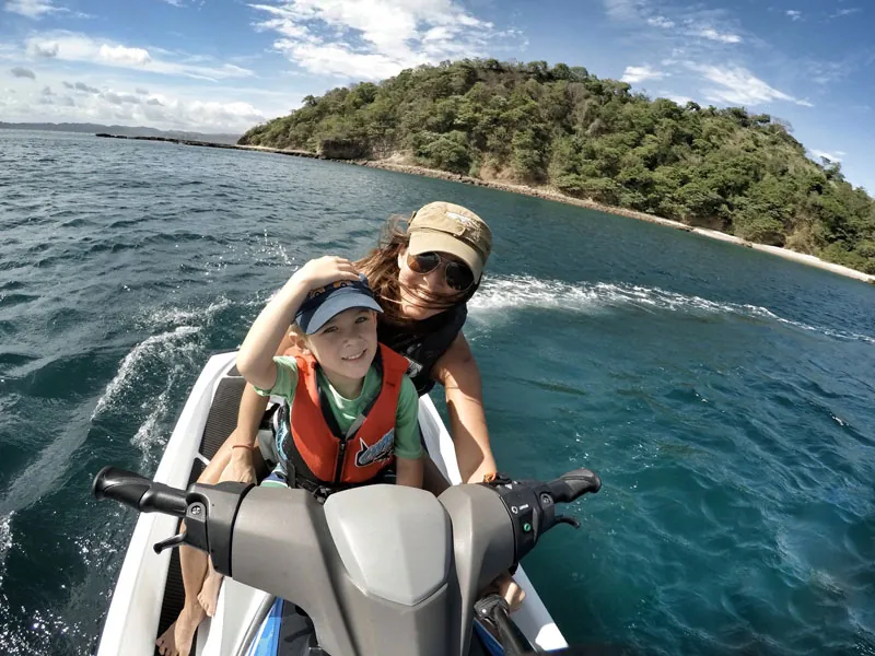 Parent and child in life vests riding a jet ski past a green island in Banderas Bay