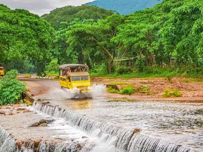 Yellow open-air Unimog truck fording a river beside a low weir, jungle hills behind