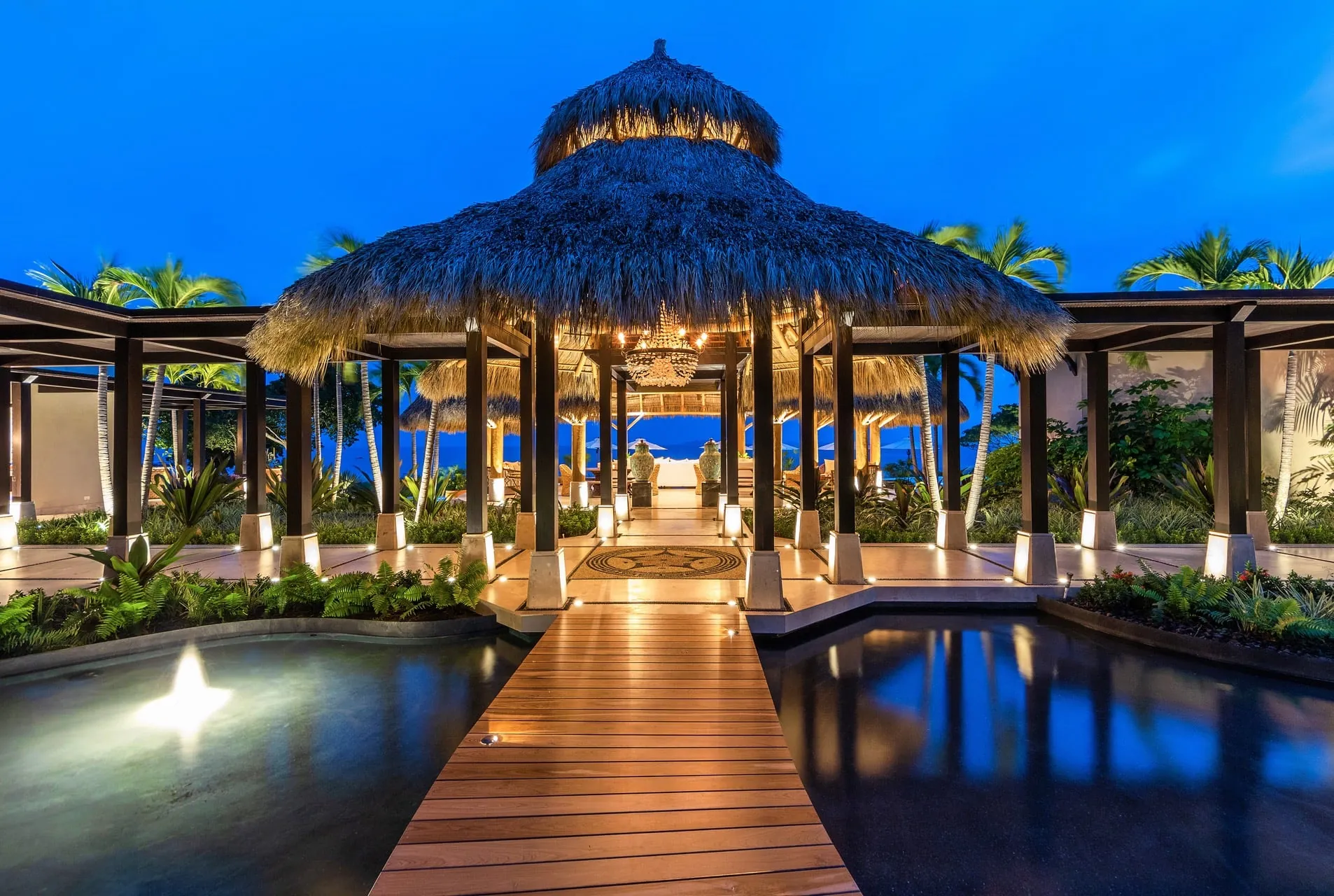 Lit wooden walkway crossing reflecting pools to a tall thatched palapa entry pavilion at blue hour, with the ocean visible beyond.