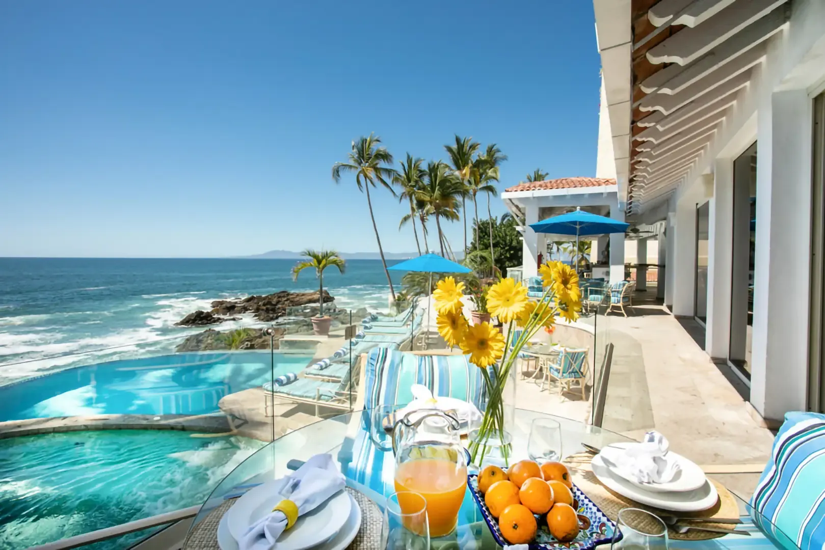 Breakfast table set with fresh orange juice, oranges and yellow gerberas on an oceanfront terrace beside an infinity pool and rocky shoreline.