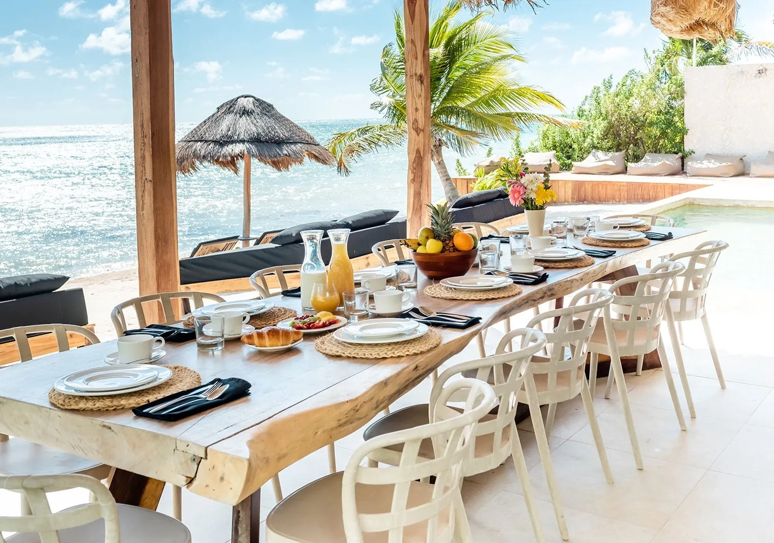 Long live-edge wood table set for breakfast with juice, fruit and flowers under a palapa facing a sunlit Caribbean beach.