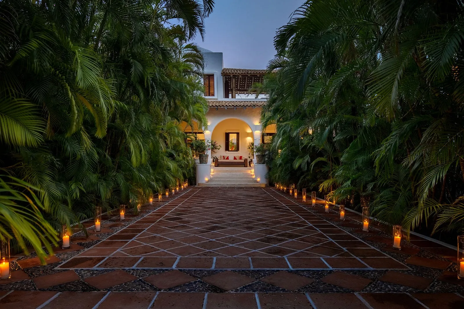 Candle-lined terracotta tile walkway through tall palms leading to a lit white villa entrance with an arched porch at dusk.