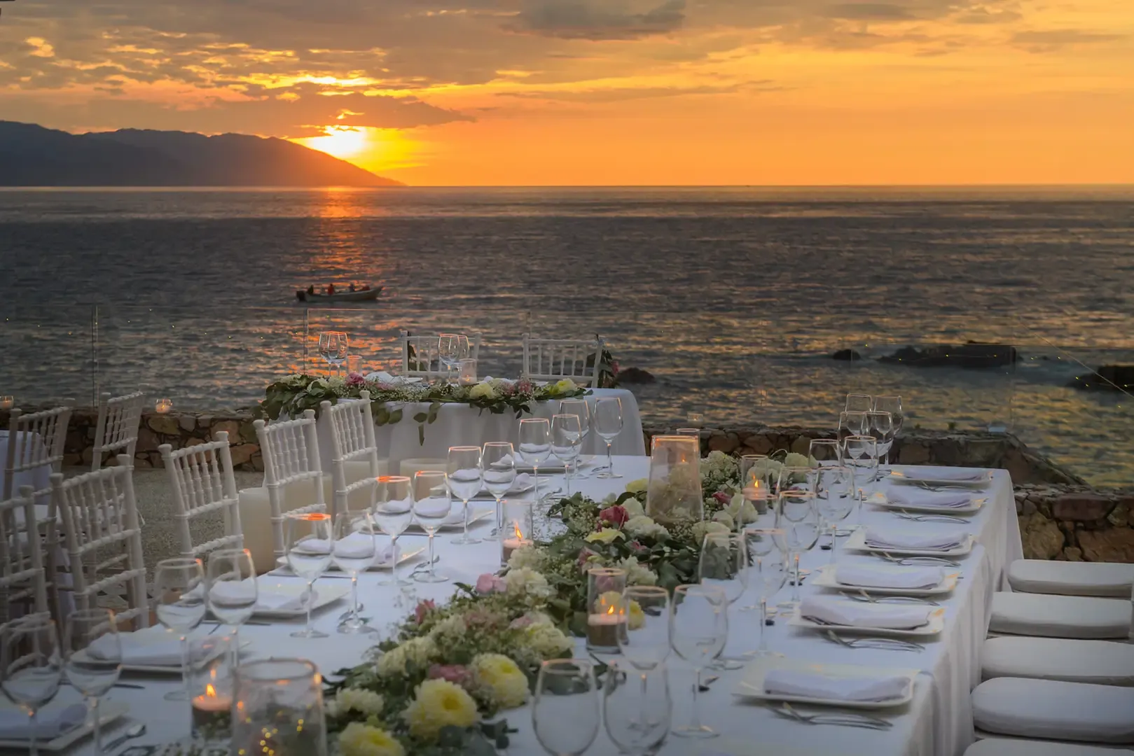 Long candlelit dinner table with white linens and floral garland set on a seaside terrace as the sun sets over the bay.