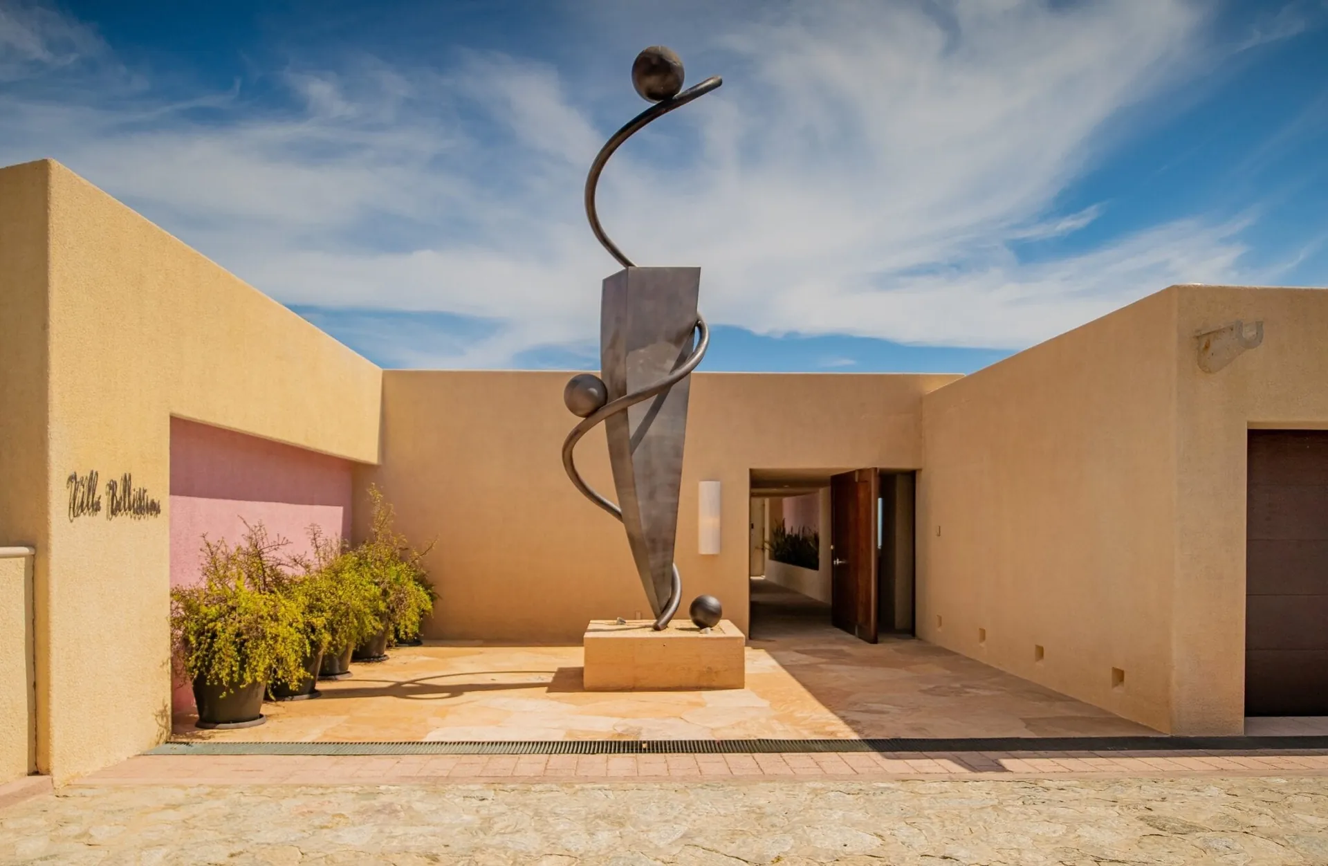 Ochre-walled entry court with a tall abstract metal sculpture and an open wooden front door under a blue sky.