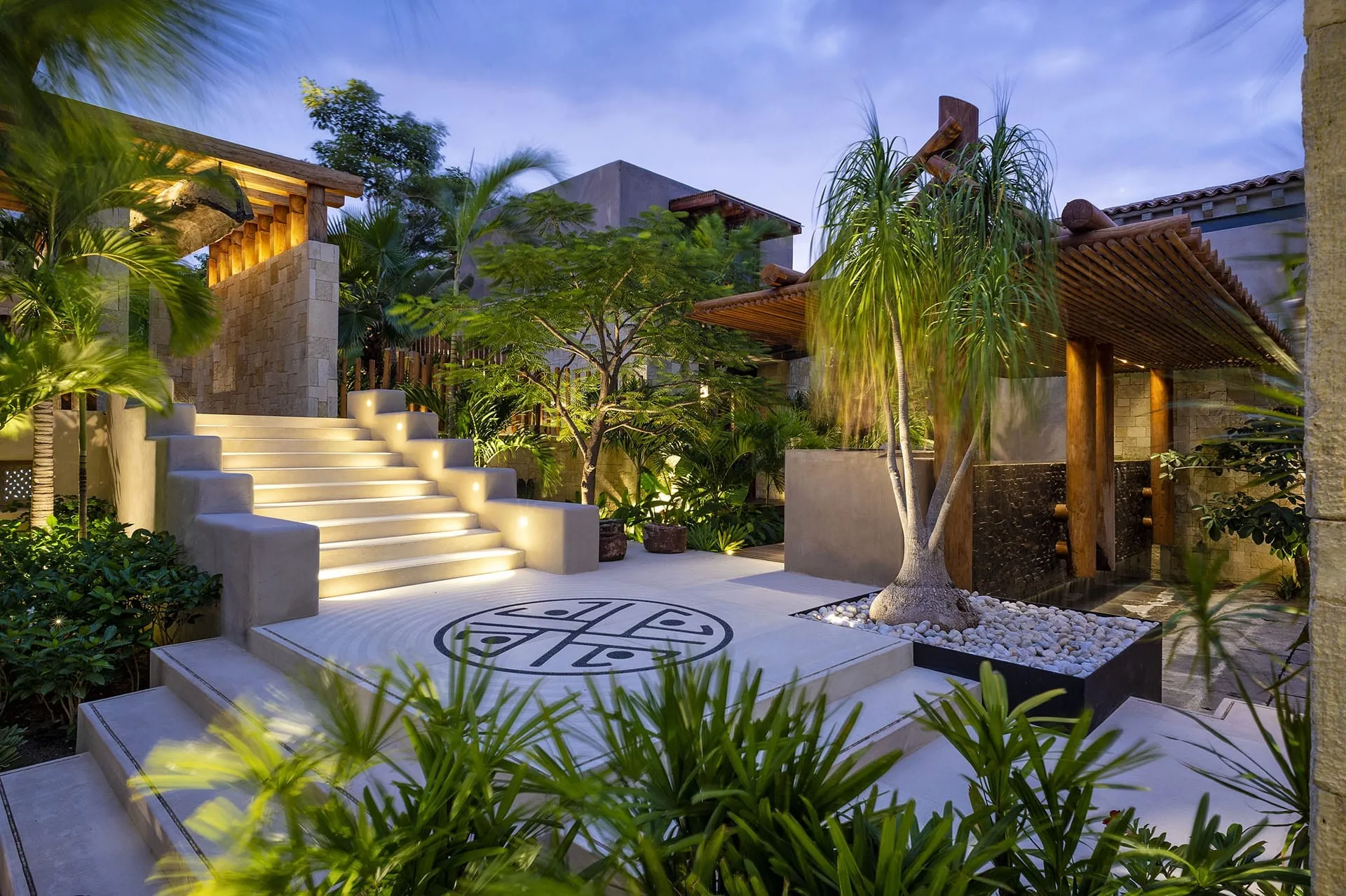 Stone entry courtyard at dusk with softly lit stairs, a mosaic medallion in the paving and tropical plants around a timber pergola.