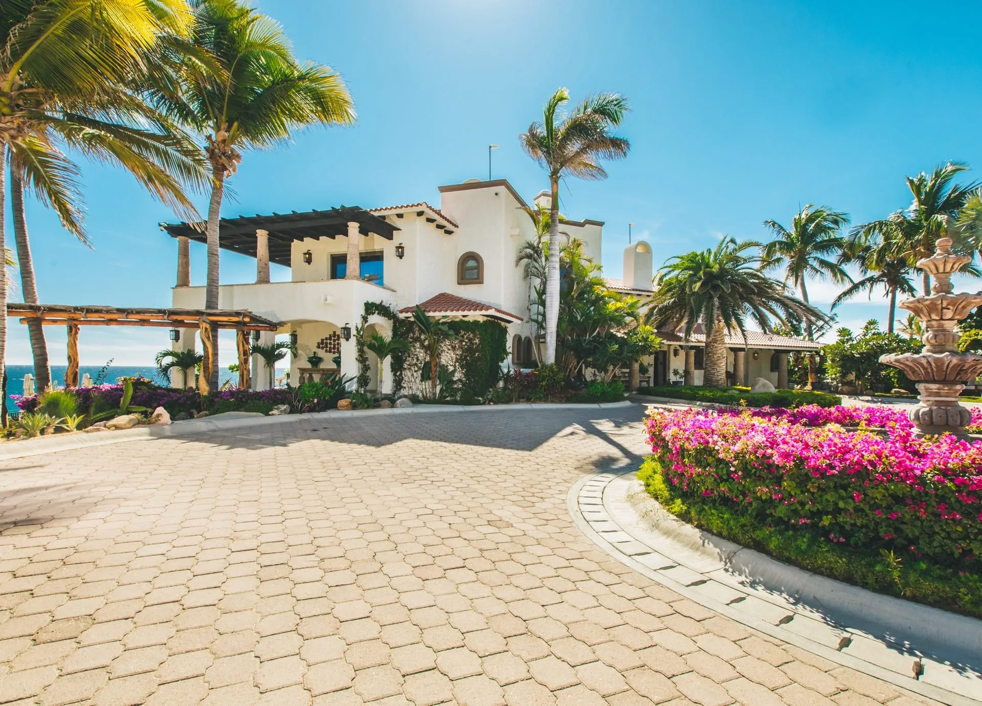 Paved circular driveway with a stone fountain and bougainvillea leading to a white Spanish-colonial villa framed by palms, ocean at left.