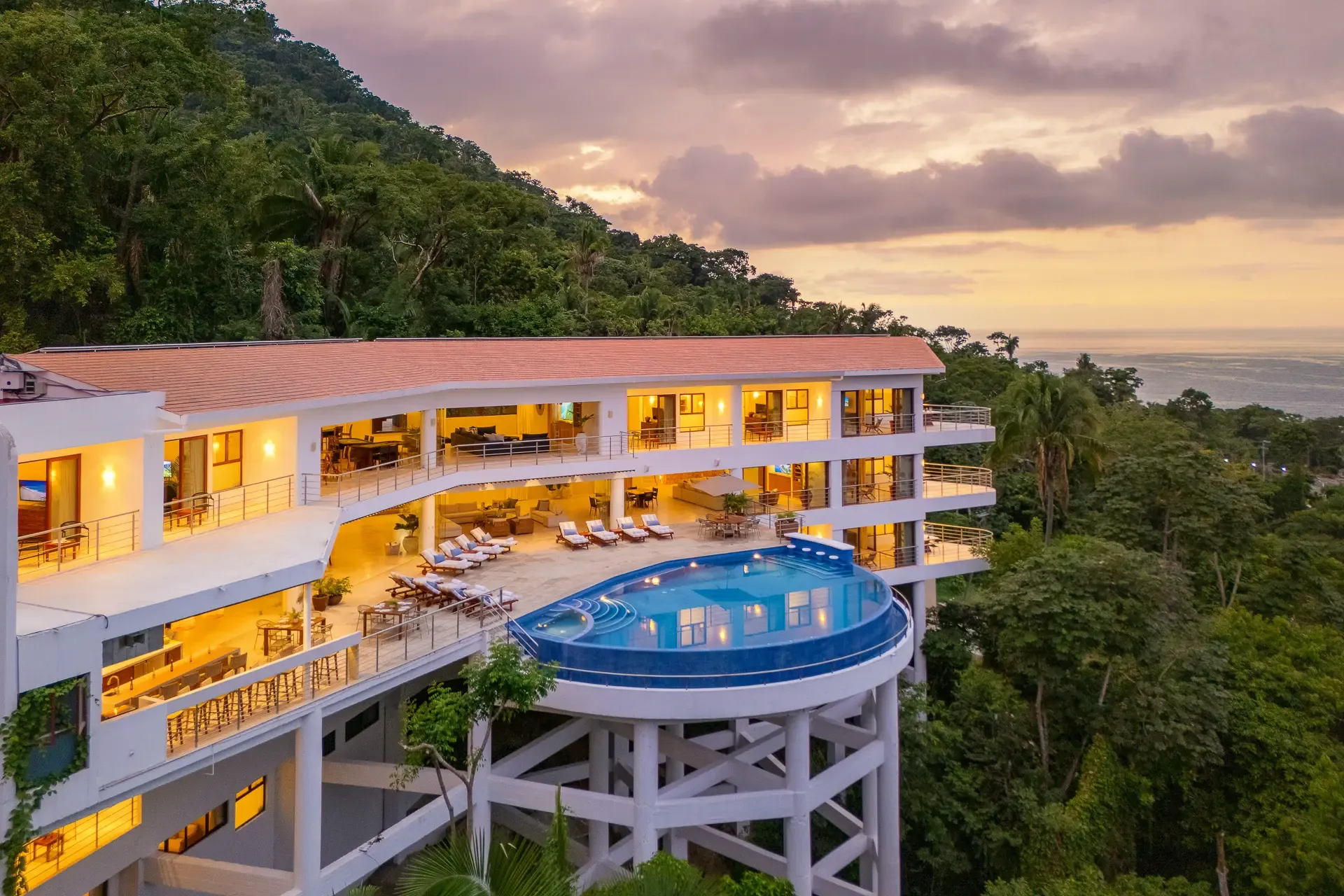 Aerial view at sunset of a multi-level white hillside villa with a round infinity pool on a stilted platform, surrounded by jungle above the bay.