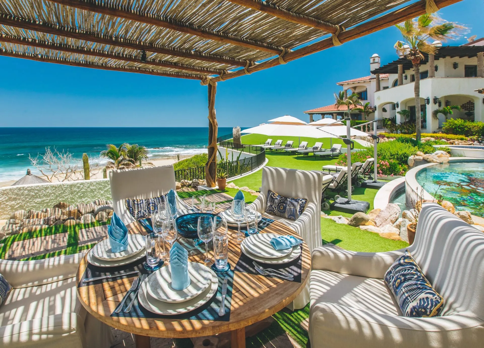 Round table set for six with blue napkins and crystal under a shade pergola, overlooking a beach, lawn and pool.