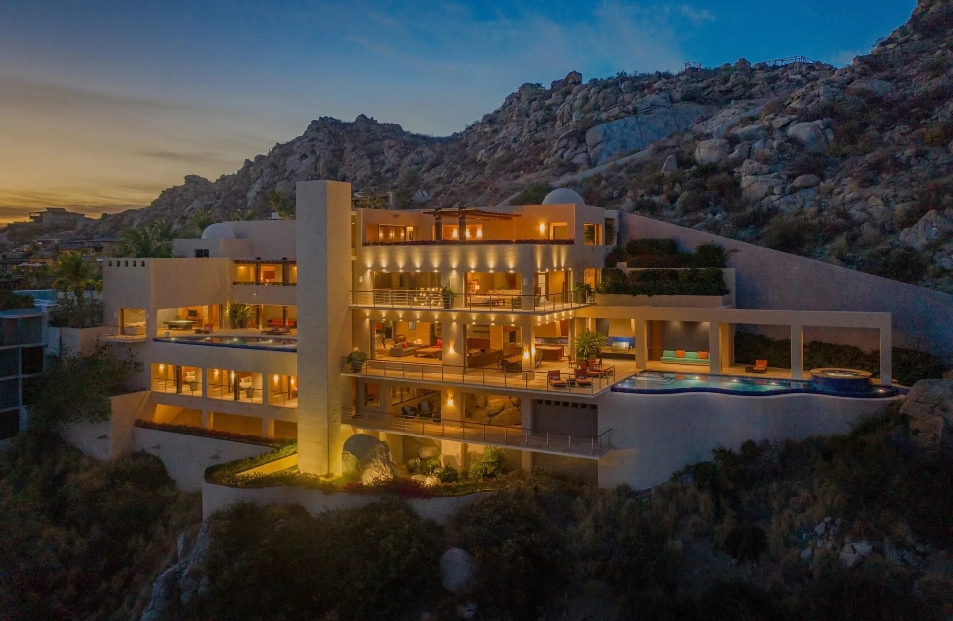 Multi-level modern villa glowing at twilight against a rocky desert hillside, with an infinity pool on the lower terrace.