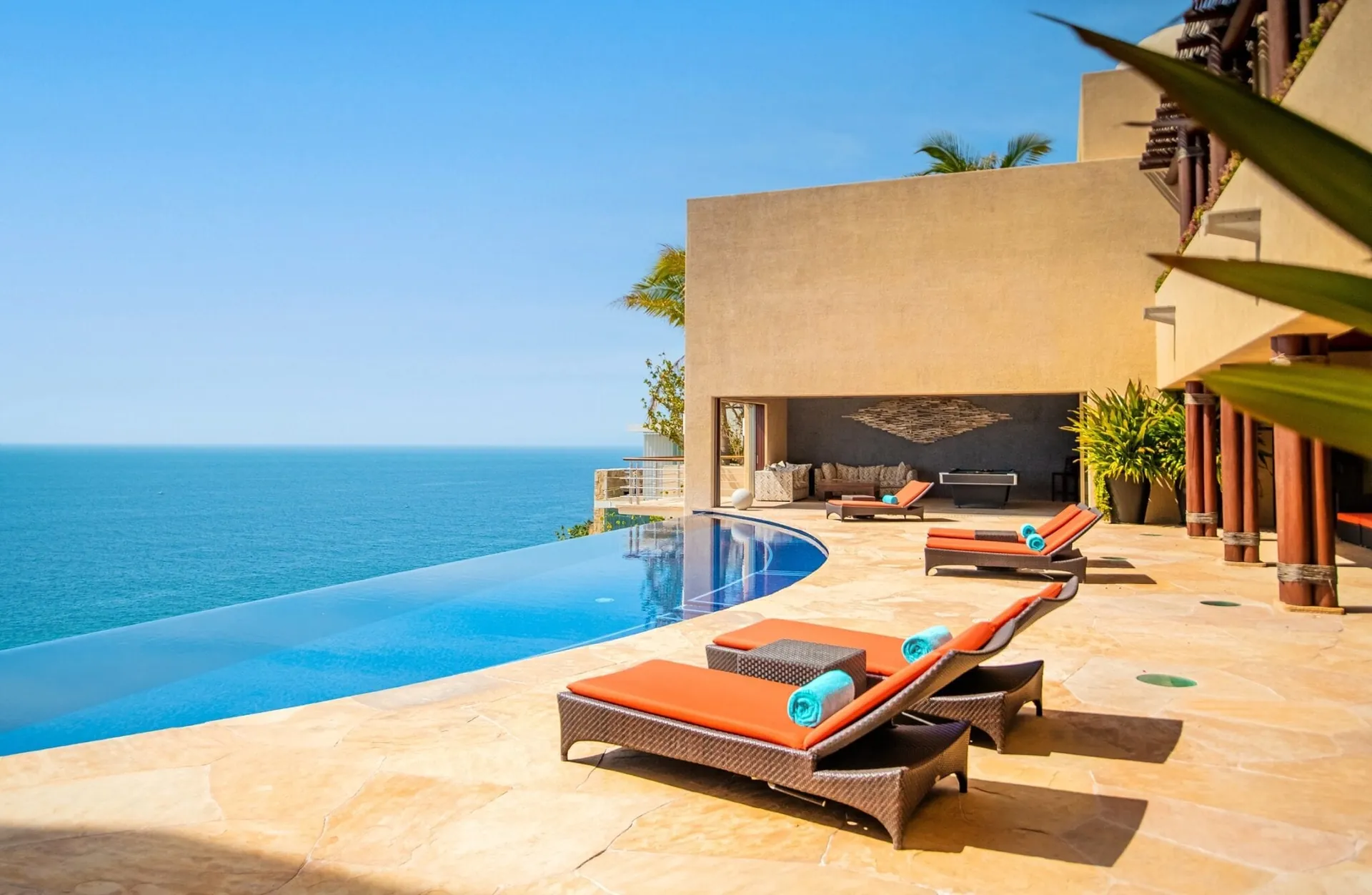 Orange-cushioned loungers on a stone pool deck beside an infinity pool that meets the open Pacific.