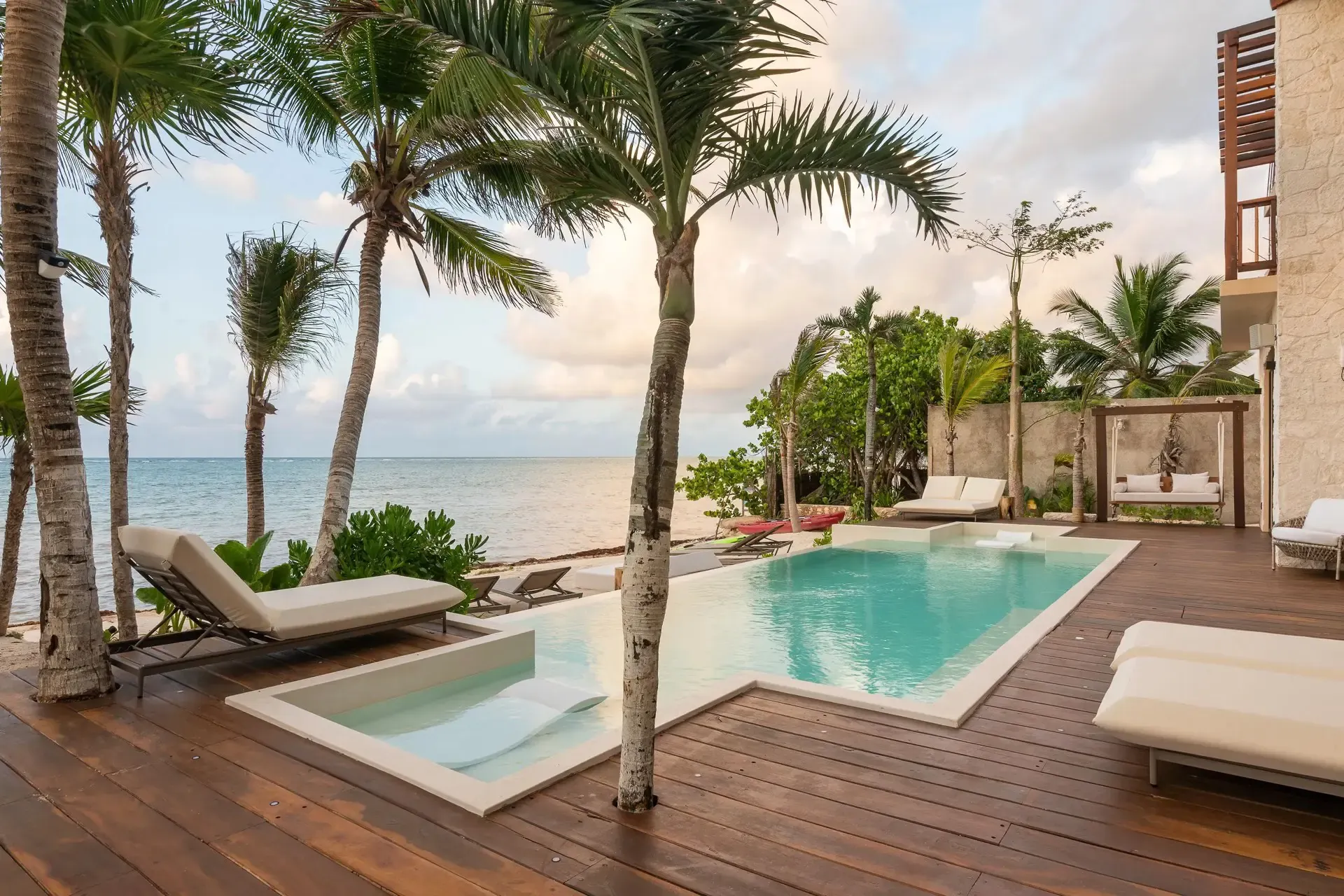 Wooden pool deck with a shallow-ledge pool, palms and cushioned loungers steps from the Caribbean shore under a soft sky.