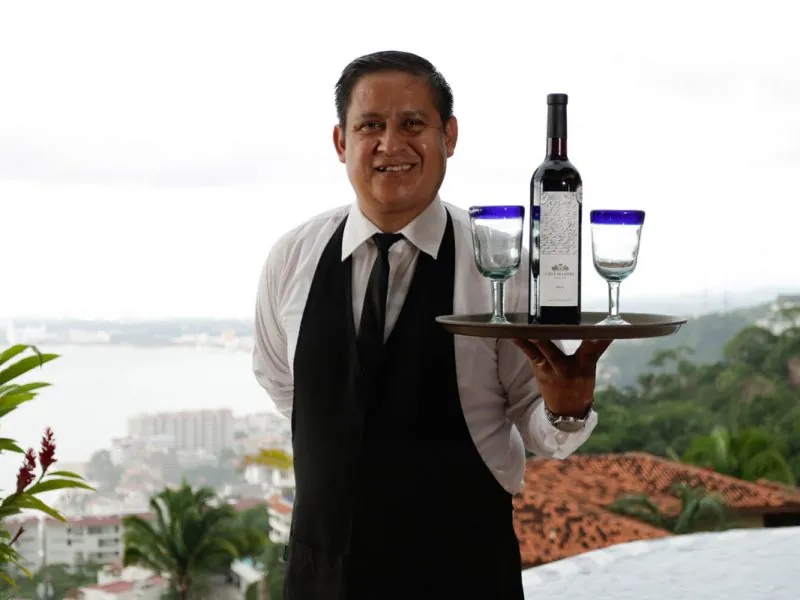 Waiter in a black vest holding a tray with wine and blue-rimmed glasses above the rooftops of Puerto Vallarta