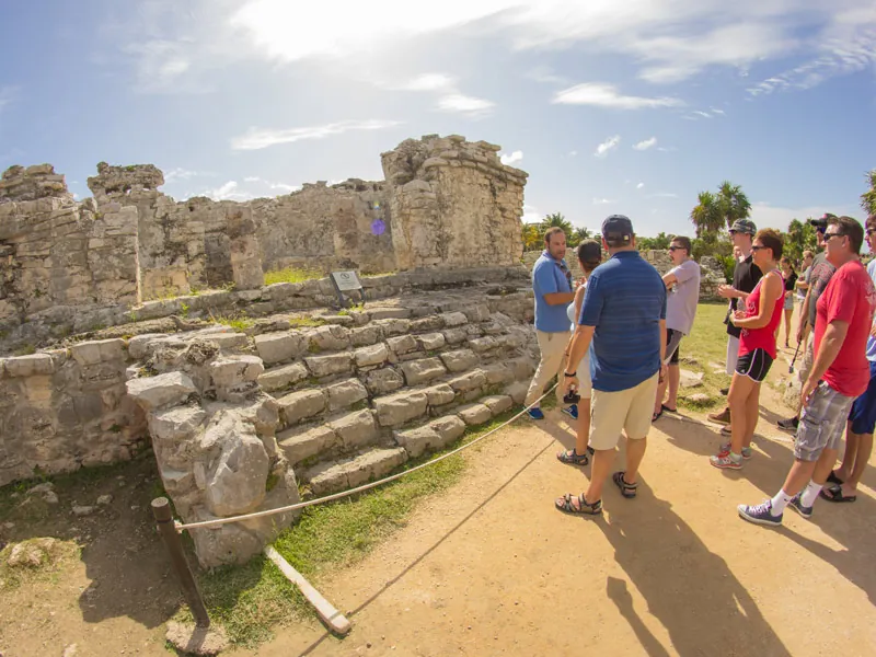 A small group of visitors listening beside low stone ruins at Tulum on a sunny day