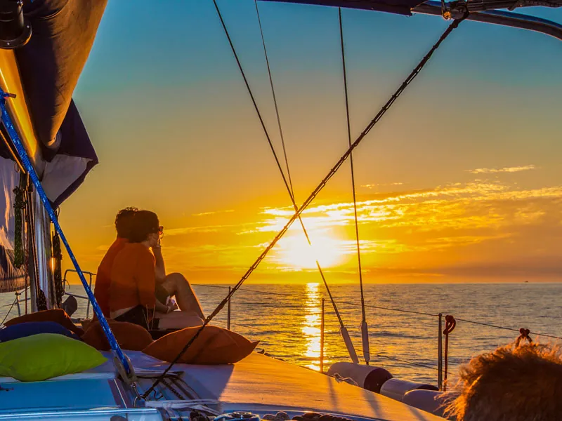 Couple sitting at the bow of a sailboat watching the sun drop to the horizon