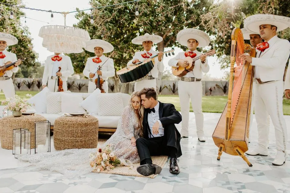 Newlyweds sitting on a terrace surrounded by a mariachi band in white charro suits with violins, guitars and a harp