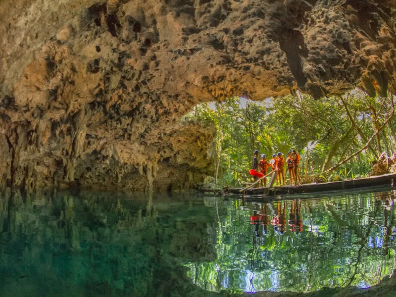 Visitors on a wooden platform at the jungle opening of a cenote cave, reflected in green water