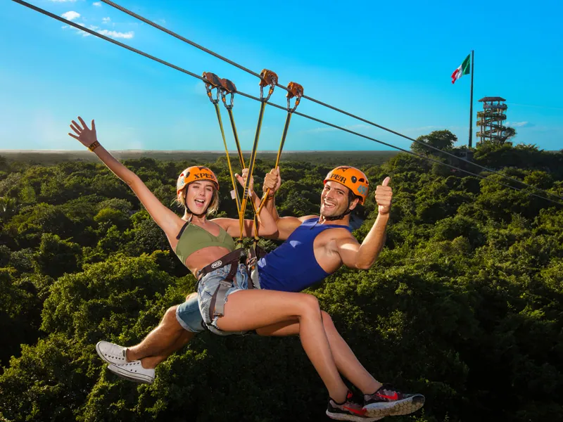 A couple in orange helmets ride a zip-line together above the jungle canopy at Xplor